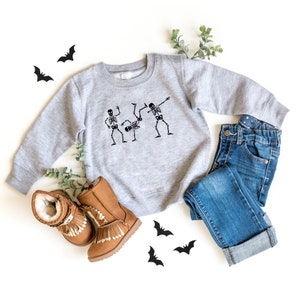 May include: A gray sweatshirt with a design of three skeletons dancing. The sweatshirt is paired with blue jeans and brown boots. There are black bats scattered around the sweatshirt.