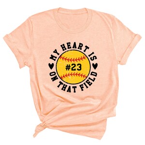 May include: Peach colored t-shirt with a black and white graphic of a softball with the text "My heart is on that field" and the number "#23".