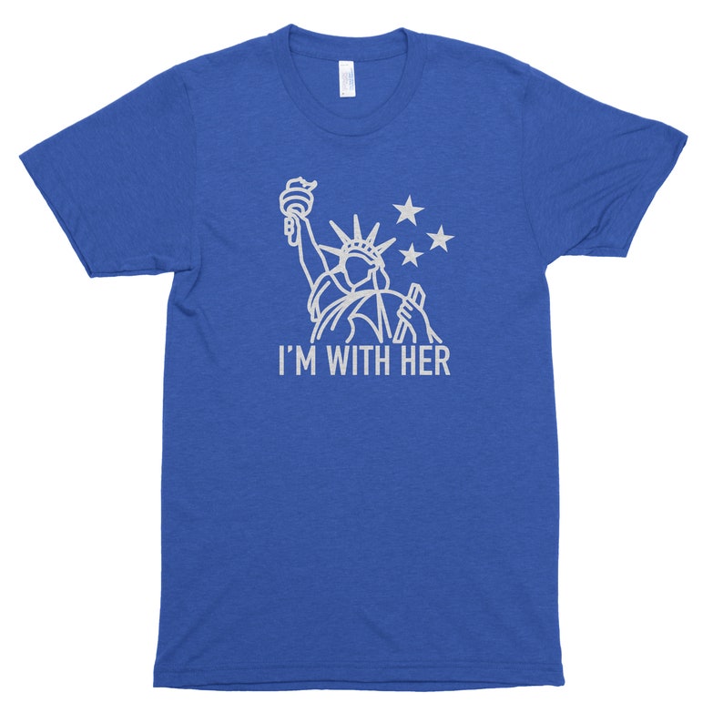 Puede incluir: Una camiseta azul real con un contorno blanco de la Estatua de la Libertad y el texto "I'M WITH HER".
