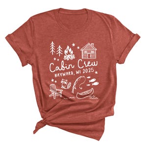 May include: A rust-colored t-shirt with a white graphic design. The design includes the text "Cabin Crew Hayward, WI 2025" along with illustrations of trees, a campfire, a cabin, a raccoon, a fishing rod, fish, a canoe, and a chair.