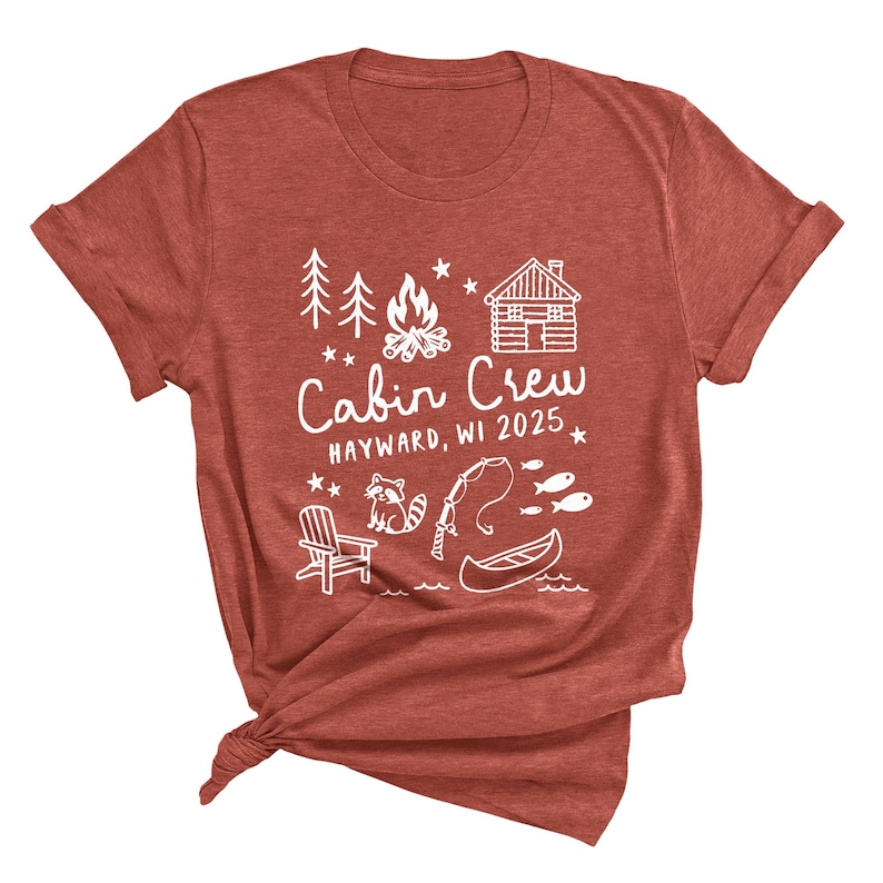 May include: A rust-colored t-shirt with a white graphic design. The design includes the text "Cabin Crew Hayward, WI 2025" along with illustrations of trees, a campfire, a cabin, a raccoon, a fishing rod, fish, a canoe, and a chair.
