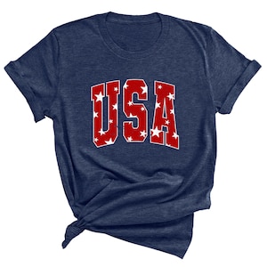 K&ouml;nnte beinhalten: Ein marineblaues T-Shirt mit einer roten "USA"-Grafik. Die Buchstaben sind mit wei&szlig;en Sternen gef&uuml;llt.