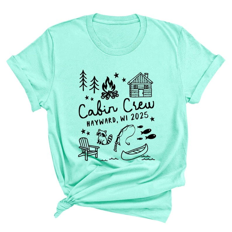 May include: A light teal t-shirt with a black graphic design. The design includes a cabin, campfire, trees, stars, a raccoon, fishing rod, fish, a canoe, and the text "Cabin Crew HAYWARD, WI 2025".