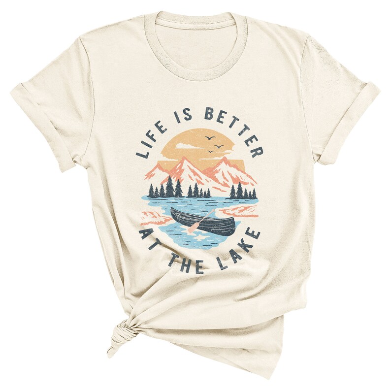 May include: A cream coloured t-shirt with a vintage-style graphic of a lake scene with mountains, trees, and a canoe. The text "Life is better at the lake" is printed in a circle around the graphic.