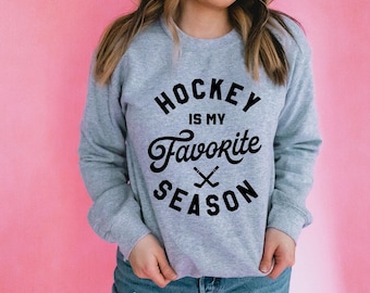 May include: A light gray crewneck sweatshirt featuring black text that reads HOCKEY IS MY FAVORITE SEASON with a graphic of two crossed hockey sticks positioned between the words Favorite and Season on the front of the garment.