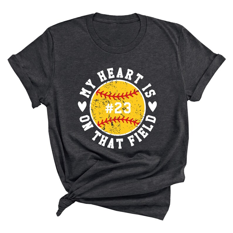 May include: A dark gray t-shirt with a white circle graphic that says "My heart is on that field" with a yellow softball with red stitching and the number 23 in the center.