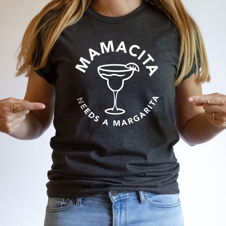 Puede incluir: Una camiseta gris oscuro con un gr&aacute;fico blanco que dice "Mamacita Needs a Margarita" y presenta un vaso de margarita con una rodaja de lima.