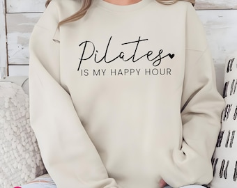 May include: A cream-colored crewneck sweatshirt featuring the black text "Pilates IS MY HAPPY HOUR" with a small heart icon at the end of the word Pilates. The garment has long sleeves and a ribbed neckline, providing a simple and clean look.