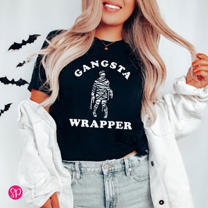 May include: Black t-shirt with the words "GANSTA WRAPPER" in white, featuring a mummy graphic. The shirt is a Halloween-themed design. Decorative black bat cutouts are in the background.
