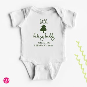 May include: White baby onesie with the text "little hiking buddy ARRIVING FEBRUARY 2026" in dark green. A dark green tree graphic is above the words "hiking buddy". The onesie has short sleeves and a round neckline.