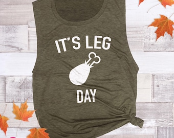 May include: A heathered olive green sleeveless tank top featuring the white text IT'S LEG DAY and a graphic of a chicken leg drumstick centred on the front. The garment has a round neckline and a relaxed fit with a knotted hem detail.