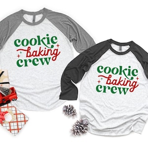May include: Two raglan baseball shirts with heather gray bodies and dark gray sleeves. The shirts feature the text "cookie baking crew" in green and red lettering with small star accents. A red toy truck and gift boxes are visible.