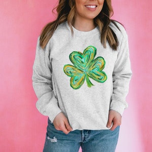 May include: A light gray sweatshirt featuring a vibrant, abstract shamrock design in shades of green, yellow, and blue. The shamrock is the focal point, set against the neutral backdrop of the sweatshirt. The sweatshirt is paired with blue jeans.