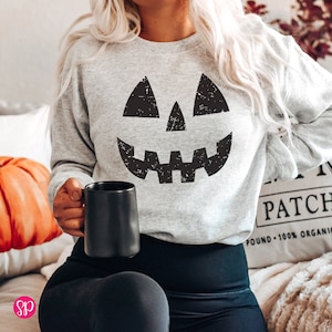 May include: A gray sweatshirt with a black distressed jack-o'-lantern face graphic. The sweatshirt is worn by a person holding a black mug.