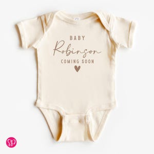 May include: Cream-colored baby onesie with the text "BABY Robinson COMING SOON" in brown lettering, along with a small heart graphic. The onesie has short sleeves and a round neckline.