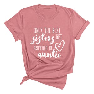 May include: A dusty rose-colored t-shirt with rolled-up sleeves. The shirt features the white text, "ONLY THE BEST sisters GET PROMOTED TO auntie" with a heart graphic. The shirt is knotted at the bottom.