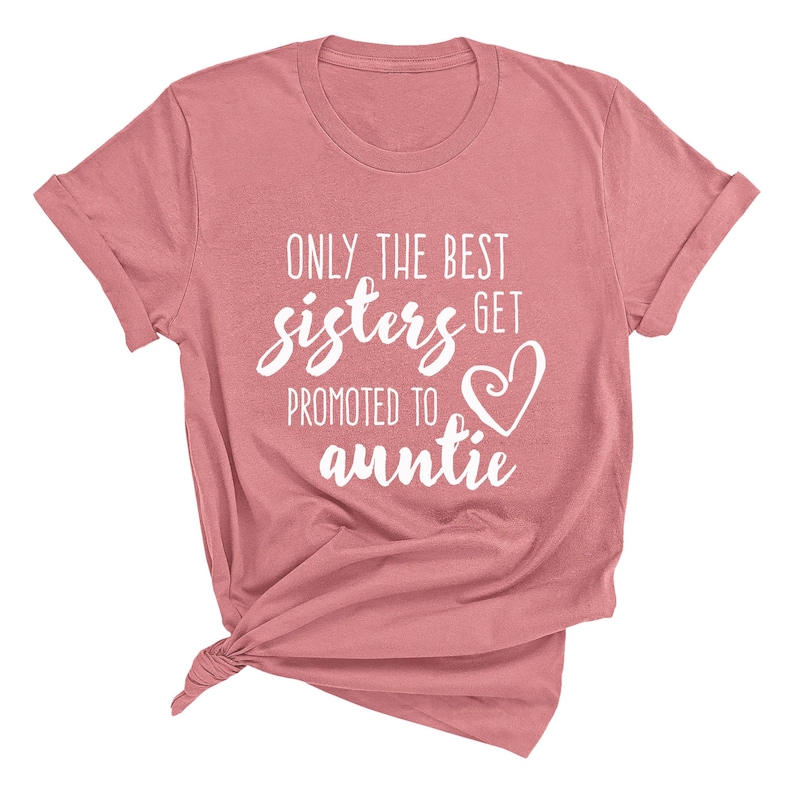May include: A dusty rose-colored t-shirt with rolled-up sleeves. The shirt features the white text, "ONLY THE BEST sisters GET PROMOTED TO auntie" with a heart graphic. The shirt is knotted at the bottom.