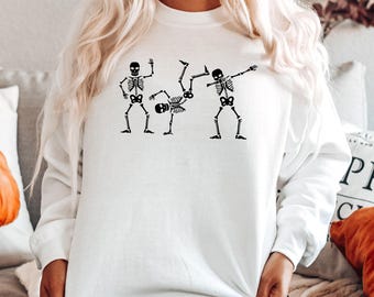 May include: A white long-sleeve sweatshirt featuring a black graphic print of three skeletons in different dance poses, including one performing a handstand and another performing a dab, centered on the chest area of the garment.