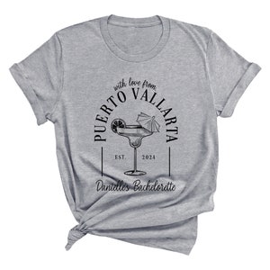May include: Gray t-shirt with black text that reads "with love from PUERTO VALLARTA EST. 2024" and "Dant&eacute;lle's Bachelorette" with a drawing of a margarita glass with a lime and an umbrella.