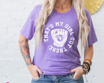 May include: A purple short-sleeve t-shirt featuring a white graphic of a baseball glove with the number 4 inside. The text surrounding the glove reads, THAT'S MY GIRL OUT THERE! The shirt is paired with blue denim jeans.