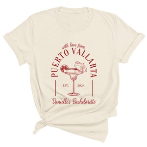 May include: A white t-shirt with a red circle design that says "Puerto Vallarta" with a margarita glass and the text "with love from" and "Danielle's Bachelorette" with the year "2024" and "EST."