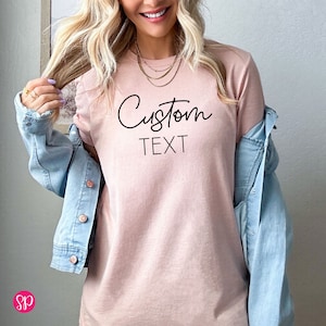 May include: A light pink t-shirt with the text "Custom Text" in black script font.