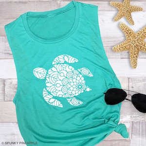 May include: A teal sleeveless tank top with a white mandala-style sea turtle design. The tank top is on a white wood background with two starfish.