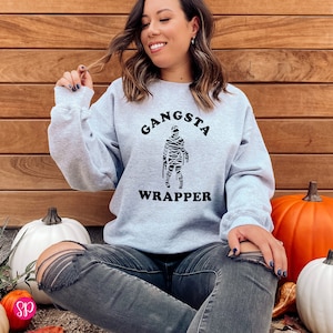 May include: A light gray sweatshirt with the words "Gangsta Wrapper" printed in black. The graphic features a mummy illustration. The sweatshirt is a crew neck style and is made of a soft material.