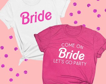 May include: A white t-shirt with the word Bride in hot pink script and a hot pink t-shirt with the white text Come on Bride Let's Go Party. Both shirts feature short sleeves and crew necklines, displayed against a light pink background.
