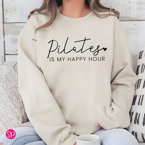May include: A cream-colored sweatshirt with the text "Pilates is my happy hour" in black lettering. A small heart symbol is next to the word "Pilates".