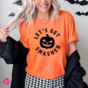 May include: Orange t-shirt with the text "LET'S GET SMASHED" surrounding a black jack-o'-lantern graphic. The shirt is a crew neck style with rolled-up sleeves. The person is wearing a black cardigan and black and white checkered pants.
