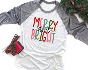 May include: A heather gray and white raglan long-sleeve shirt featuring the text MERRY and BRIGHT in red, green, and pink lettering. The shirt has a round neckline and gray sleeves that contrast with the white heathered torso section.