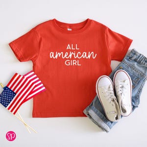 May include: Red short-sleeved t-shirt with the text "ALL american GIRL" in white. The shirt is paired with blue jeans and white sneakers.