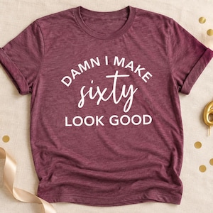 May include: A maroon short-sleeved t-shirt with white text printed on the front that reads DAMN I MAKE sixty LOOK GOOD. The shirt features a crew neckline and a heathered fabric texture, presented as a flat lay garment for a birthday celebration.