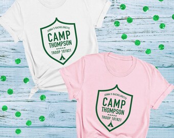 May include: A white t-shirt and a light pink t-shirt, each featuring a dark green shield graphic with the text "JENNA'S BACHELORETTE CAMP THOMPSON TROOP 101821" and a small tent icon. Both shirts have short sleeves and a crew neckline.