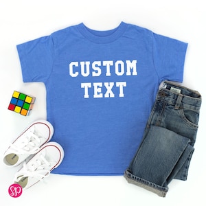 May include: A blue t-shirt with the text "CUSTOM TEXT" in white block letters. The shirt is paired with a pair of blue jeans, white sneakers, and a Rubik's Cube. The items are arranged on a white surface.