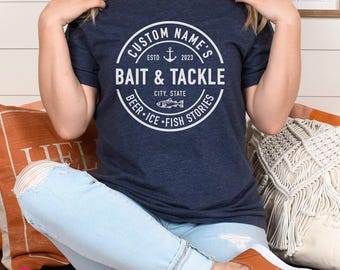 Op de afbeelding: Een marineblauw t-shirt met korte mouwen en een witte cirkelvormige afbeelding met een ankerpictogram en de tekst: CUSTOM NAME'S, ESTD 2023, BAIT & TACKLE, CITY, STATE, BEER, ICE, FISH STORIES, en een kleine visillustratie in het midden.