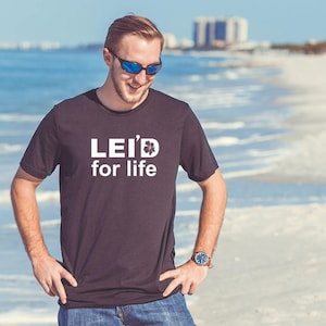 May include: Black t-shirt with the text "LEI'D for life" in white, featuring a flower graphic. The shirt is worn by a person on a beach, with sunglasses and a watch.