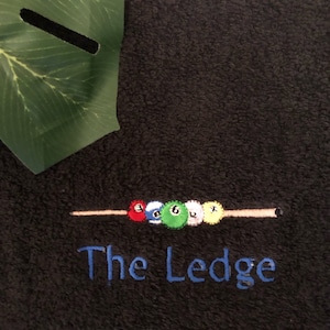 May include: Dark grey towel featuring embroidered billiard balls on a cue. The text "The Ledge" is embroidered in blue. A green leaf is in the upper left corner.