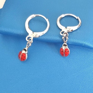 May include: A pair of silver-toned hoop earrings featuring dangling ladybird charms. Each charm is painted with a bright red enamel finish and accented with small black dots, suspended from a circular hinged hoop mechanism.