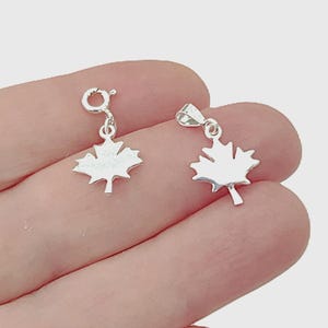 May include: Two silver-coloured maple leaf charms. One charm features a circular spring ring clasp, while the other charm is attached to a triangular bail. Both maple leaf pendants have a smooth, reflective metallic finish and a distinct leaf shape.