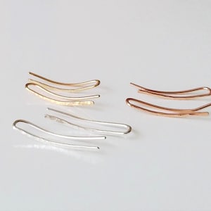 May include: Three pairs of ear climber earrings in gold, silver, and rose gold tones. Each earring features a curved, elongated wire design with a subtle hammered texture along the bottom bar, creating a sleek and minimalist look for the ears.