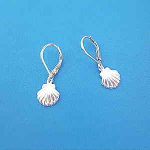 Peut inclure: Une paire de boucles d'oreilles pendantes argentées avec une petite breloque en forme de coquillage texturé suspendue à une attache dormeuse sécurisée. La surface métallique des coquillages reflète la lumière, soulignant les stries.