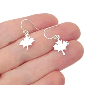 May include: A pair of silver-toned drop earrings featuring small, flat maple leaf charms. Each charm is suspended from a simple, curved silver-toned ear wire hook. The earrings have a polished, reflective surface and a clean, symmetrical design.
