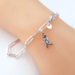 Peut inclure: Un bracelet à chaîne argentée composé de maillons rectangulaires allongés et d'une breloque unique en forme de petit personnage en costume. Le bracelet est doté d'un fermoir mousqueton et d'une petite étiquette ronde près de la fermeture.