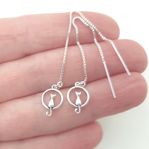 May include: A pair of silver-toned threader earrings featuring a delicate box chain that suspends a circular charm. Each charm contains a small, minimalist cat silhouette sitting inside the ring. The earrings have a polished, metallic finish.