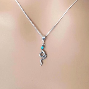 May include: A silver-toned snake pendant necklace featuring a small, bright turquoise-coloured stone set at the top of the snake's head. The pendant hangs from a thin, delicate silver-toned chain that rests against the skin in a simple, elegant display.