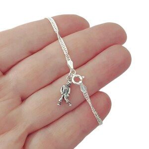 Peut inclure: Un bracelet en chaîne argentée doté d'un petit pendentif détaillé représentant une silhouette en costume. Le bracelet comprend un fermoir à ressort circulaire et un maillon tressé délicat, fabriqué en métal argenté poli et brillant.