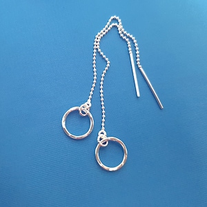 Peut inclure: Boucles d'oreilles pendantes en argent avec deux petits cercles texturés martelés. Les boucles d'oreilles ont une fine chaîne de petites perles et une barre droite qui se glisse dans l'oreille. Sur fond bleu uni.