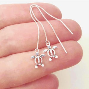 Peut inclure: Une paire de boucles d'oreilles traversantes en argent avec des breloques en forme de tortue de mer. Chaque boucle se compose d'une fine chaîne maille carrée qui passe à travers l'oreille, terminée par un pendentif tortue en argent.